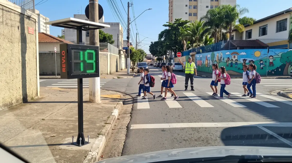 Vista de dentro de um carro: lombada eletrônica na calçada mostrando 19 em LED verde, com crianças atravessando na faixa em frente à escola